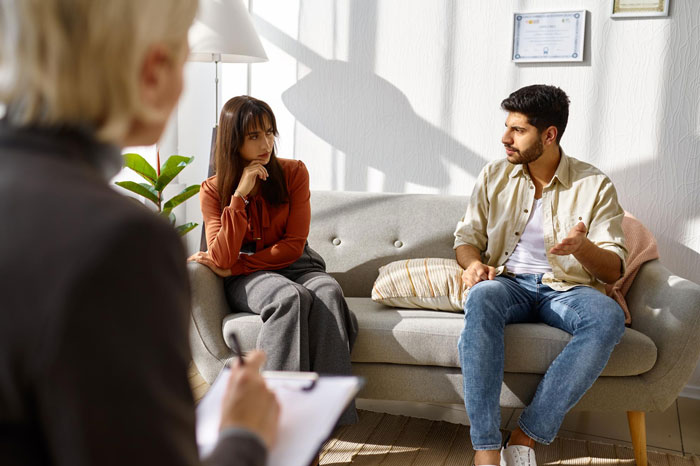 Couple in therapy discussing husband’s baby-making plan with friend while wife looks visibly upset and uncomfortable.