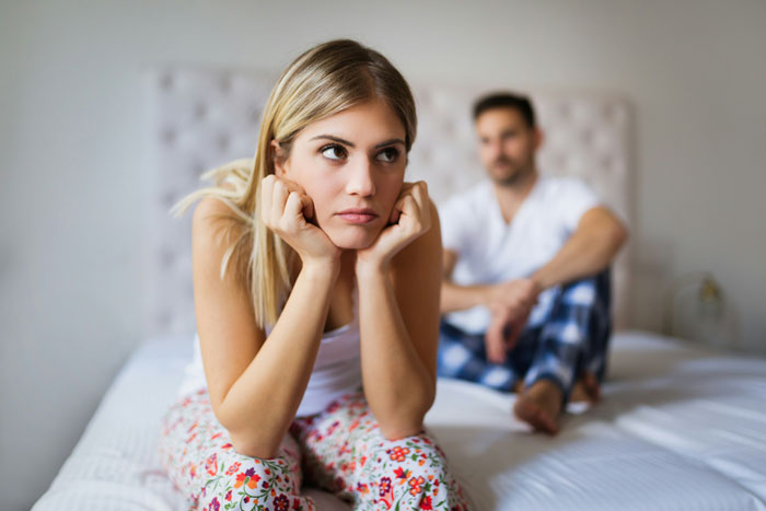 Woman looking upset and thoughtful on bed while husband in pajamas shows lack of consideration and thoughtfulness in bedroom. Woman looking upset and thoughtful on bed while husband in pajamas shows lack of consideration and thoughtfulness in bedroom.
