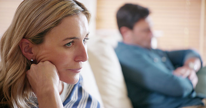 Worried wife sits on couch looking upset while jobless spouse relaxes in background draining emergency fund for World Cup