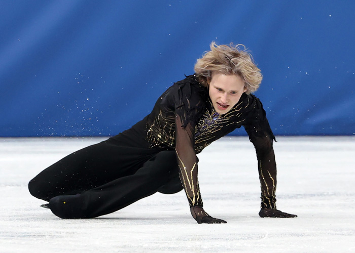 Ilia Malinin in a black and gold outfit falling on the ice during a figure skating performance at the Olympics.