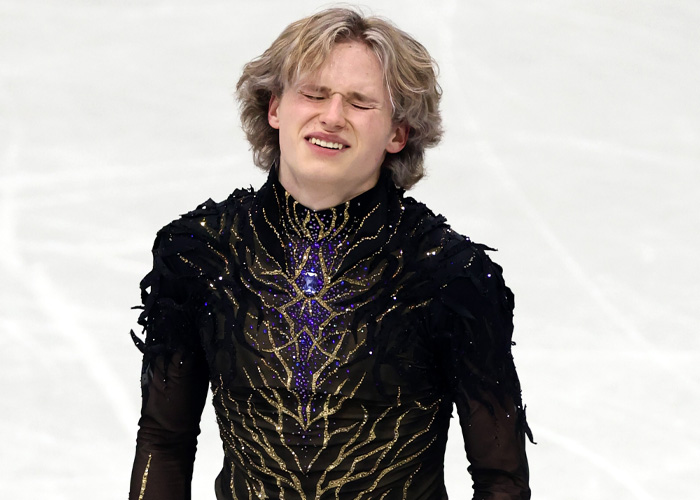 Ilia Malinin in a black costume showing a pained expression during a figure skating Olympic performance.