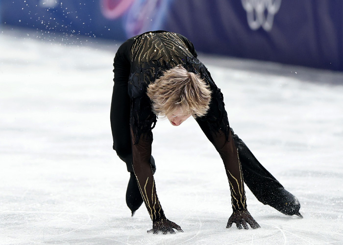 Ilia Malinin performing a figure skating routine on ice wearing a black costume during Olympic competition.