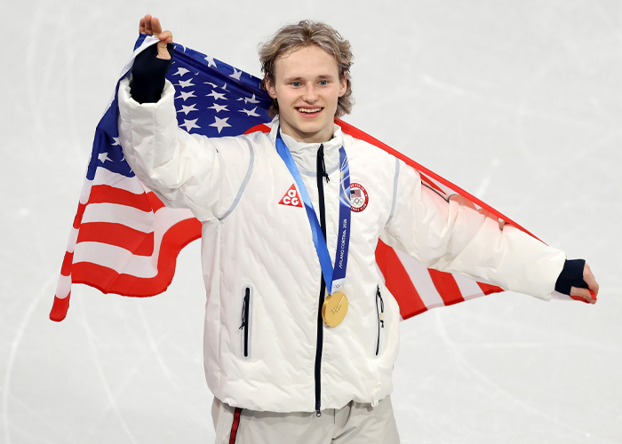 Ilia Malinin celebrating with a gold medal, draped in an American flag during an Olympic event.
