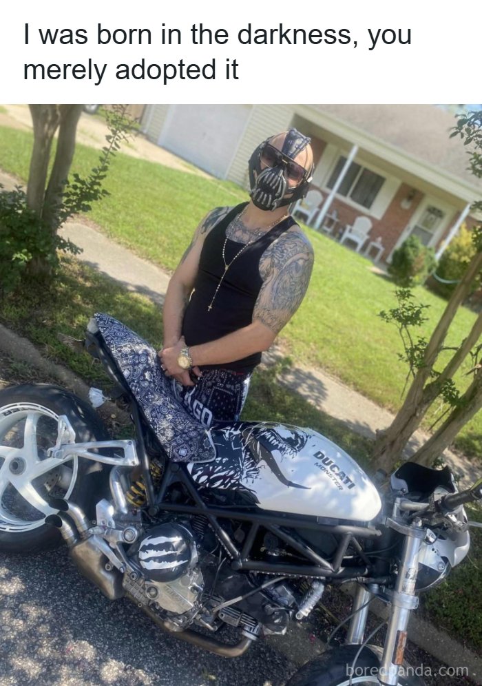 Man wearing a Bane mask and sunglasses posing with a customized Ducati Monster motorcycle outdoors.