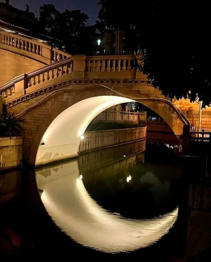 Nighttime photo of a stone bridge with illuminated arch perfectly reflecting in water creating a reality glitch illusion.
