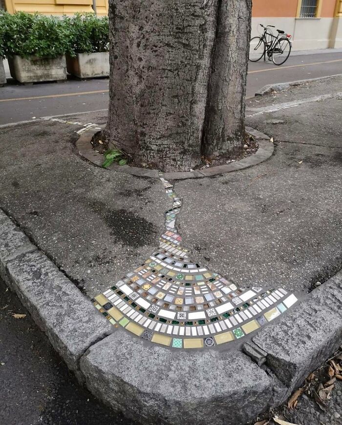 Tree trunk with mosaic tile design filling a crack in the pavement, creating a reality glitched illusion on the sidewalk.