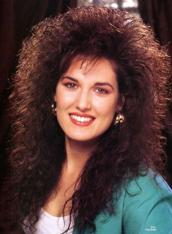 Woman with iconic 80s hairdo featuring voluminous curly hair and big volume, smiling and wearing gold hoop earrings.