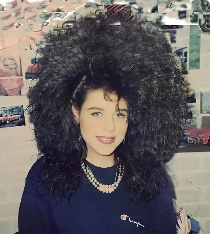 Young woman with iconic 80s hairdos featuring voluminous, curly hair and a layered necklace, wearing a dark Champion sweatshirt.