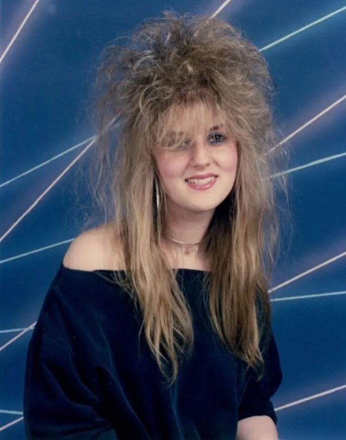 Young woman with an iconic 80s hairdo featuring voluminous teased hair and an off-the-shoulder black top against a blue background.