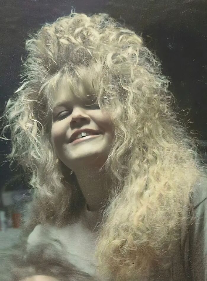 Woman with an iconic 80s hairdo featuring voluminous, curly blonde hair smiling in a dimly lit setting.