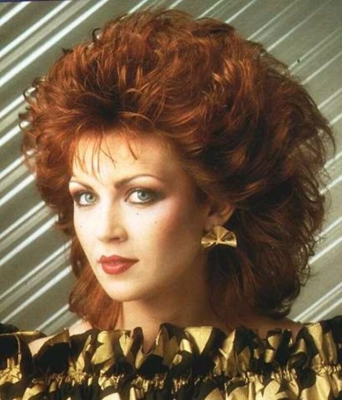 Woman with voluminous red hair styled in an iconic 80s hairdo, wearing gold earrings and bold makeup.