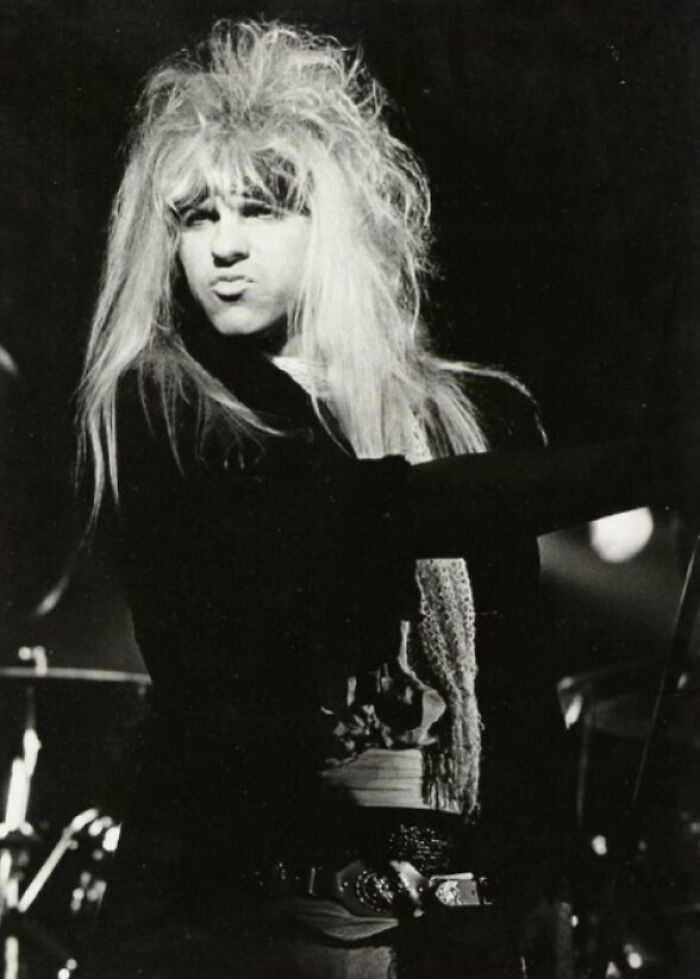 Male rock singer with iconic 80s hairdo featuring voluminous, teased long blonde hair on stage during a live performance.