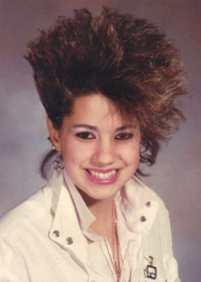 Teen girl with a voluminous 80s hairdo, wearing pink makeup and a white jacket, smiling for a portrait photo.