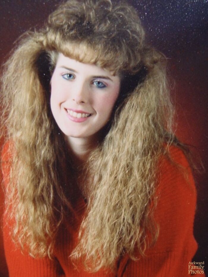 Young woman with voluminous crimped hair styled in an iconic 80s hairdo, wearing a red sweater and smiling.