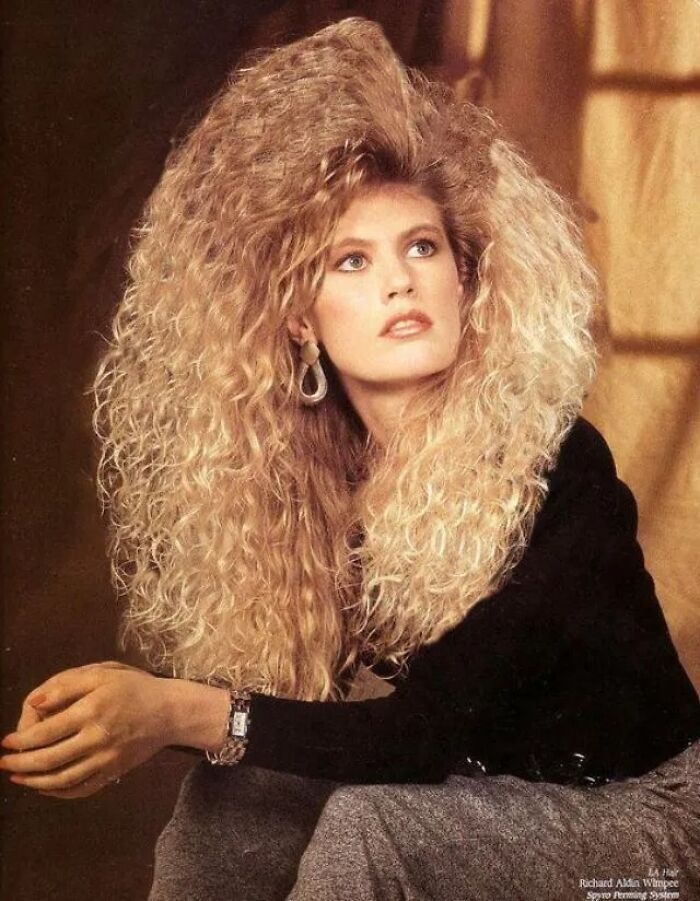 Woman with voluminous curly 80s hairdo wearing a black top and grey pants, posing with arms crossed in a vintage setting.