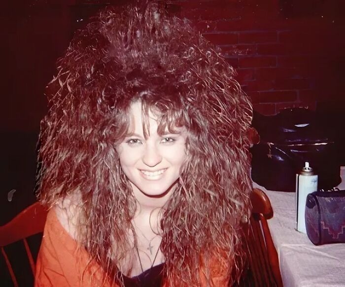 Woman with an iconic 80s hairdo featuring voluminous, teased curls and a bright smile in an indoor setting.