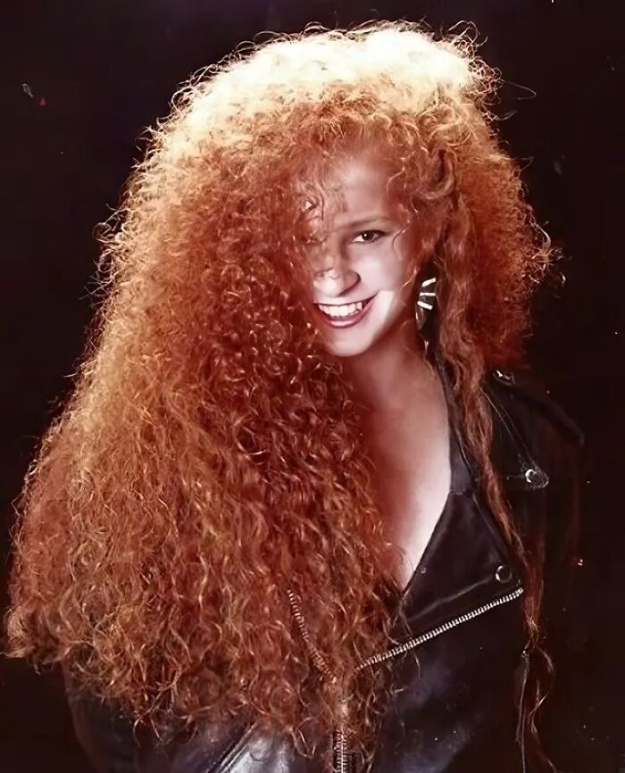 Smiling woman with voluminous curly red hair in an iconic 80s hairdo, wearing a black leather jacket against a dark background.