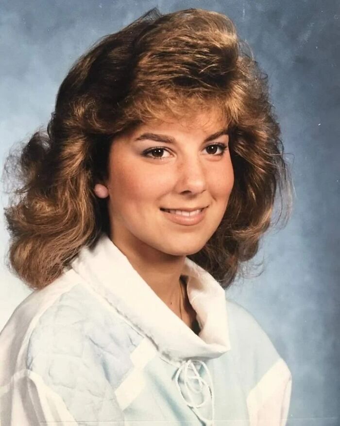 Teen girl with an iconic 80s hairdo featuring voluminous, layered waves and a side-swept style, smiling in portrait.