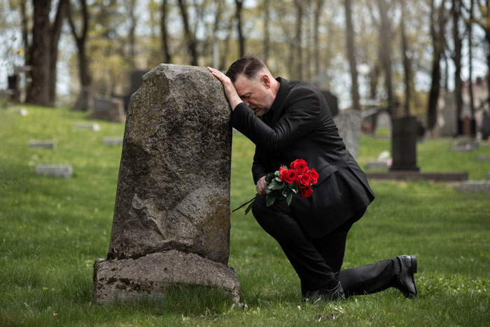 Man in black suit kneeling at late wife's grave holding red roses during a private family visit in a quiet cemetery. Man in black suit kneeling at late wife's grave holding red roses during a private family visit in a quiet cemetery.