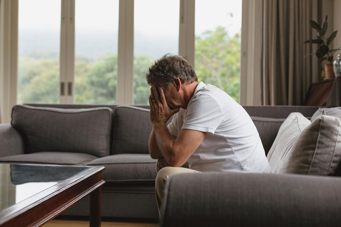 Man sitting on couch with hands covering face, reflecting on family visits to late wife’s grave and private tradition. Man sitting on couch with hands covering face, reflecting on family visits to late wife’s grave and private tradition.