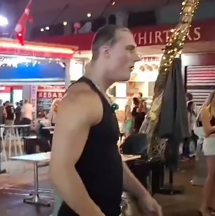 Man in black tank top reacting in crowded street scene after influencer wig snatched by stranger, capturing public humiliation moment.