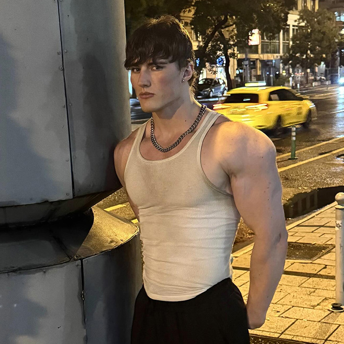 Young influencer with muscular arms wearing a beige tank top and chain necklace standing on a city street at night.
