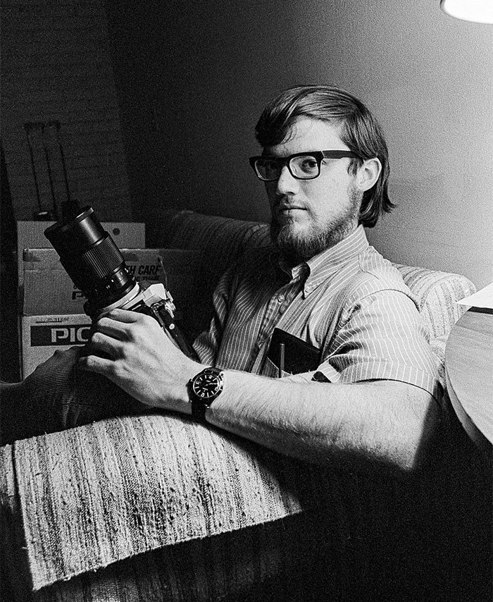 Young man with glasses holding a camera sitting on a couch in a black and white vintage photo representing hidden history moments.