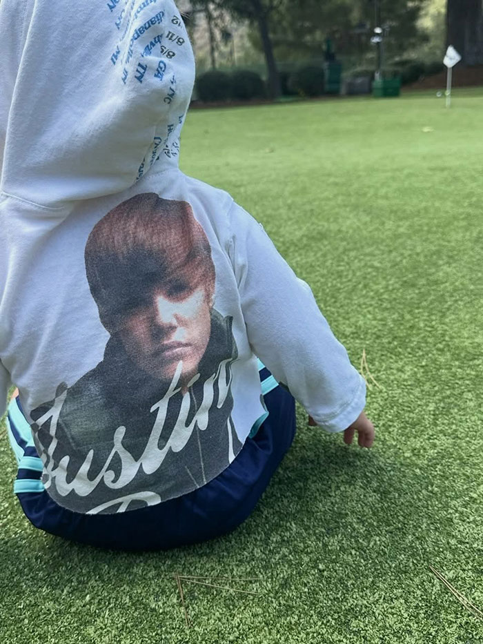 Toddler sitting on grass wearing a hoodie with a printed image, related to pathological liar and Hailey Bieber's son claim.