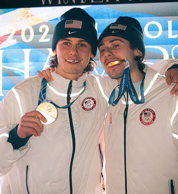 US Olympics hero Jack Hughes and teammate celebrating with gold medals wearing white team jackets and black beanies. US Olympics hero Jack Hughes and teammate celebrating with gold medals wearing white team jackets and black beanies.