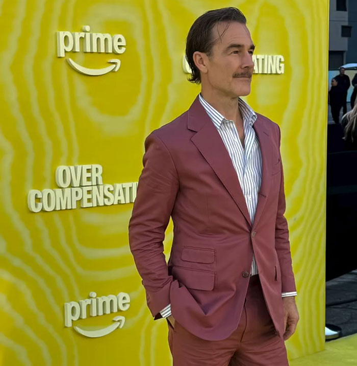 James Van Der Beek in a maroon suit at a Prime event, posing against a yellow background with Over Compensation text.