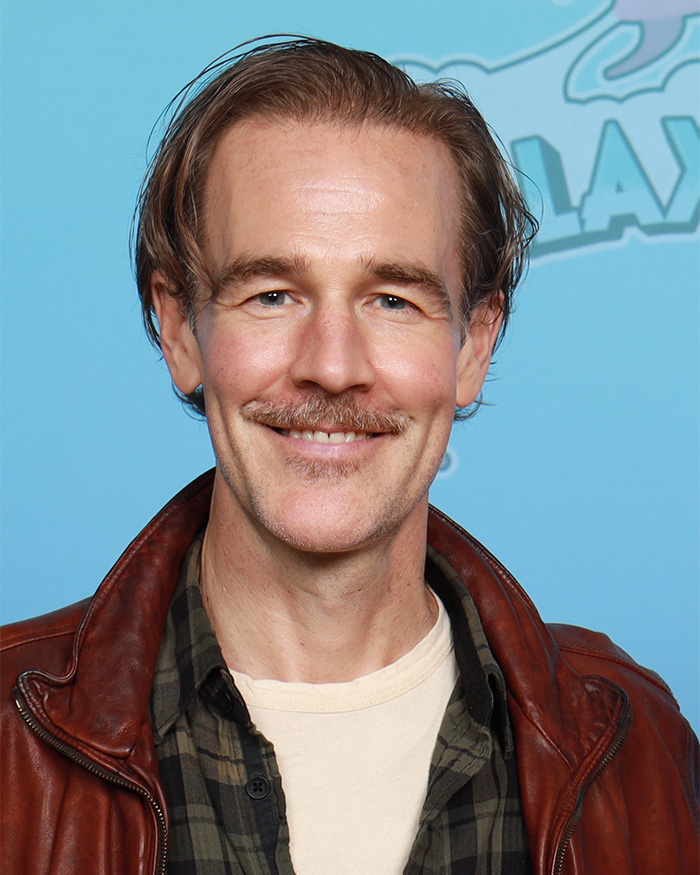 James Van Der Beek smiling, wearing a brown leather jacket and checkered shirt against a light blue background.