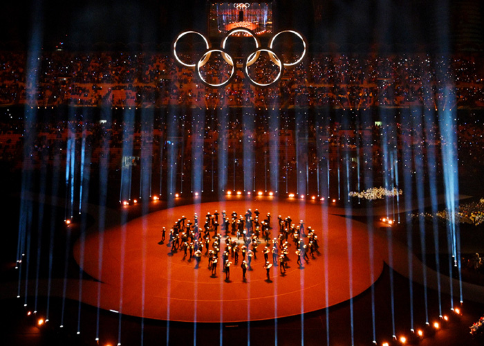 Olympic rings illuminated above performers at Winter Olympics inauguration ceremony with audience and light beams surrounding stage.
