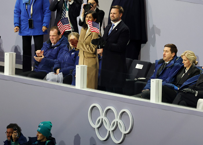 JD Vance at Winter Olympics inauguration ceremony with attendees holding American flags and Olympic rings visible below.