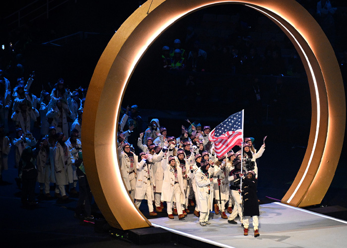 Winter Olympics athletes in white coats entering the stadium under a large golden arch during the inauguration ceremony.