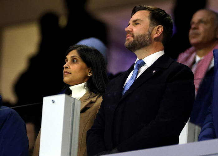 JD Vance at Winter Olympics inauguration ceremony, dressed formally, standing among other attendees with serious expressions.