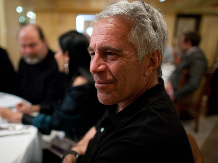 Middle-aged man with gray hair sitting at a table in a dimly lit room amid surging royal scrutiny.