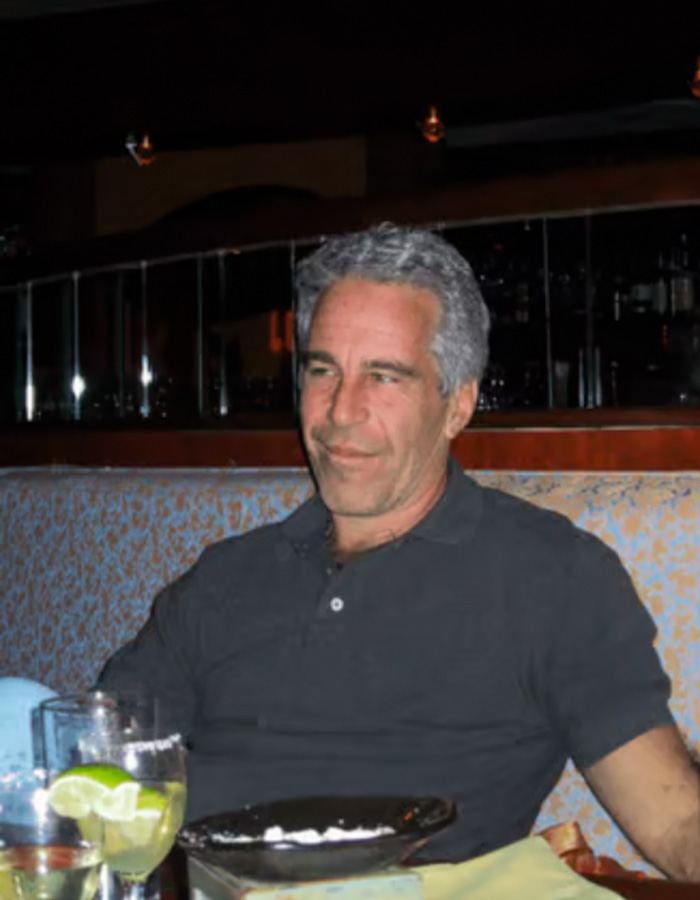 Man with gray hair wearing a dark shirt sitting at a restaurant booth with drinks and a plate nearby, Epstein obsession revealed.