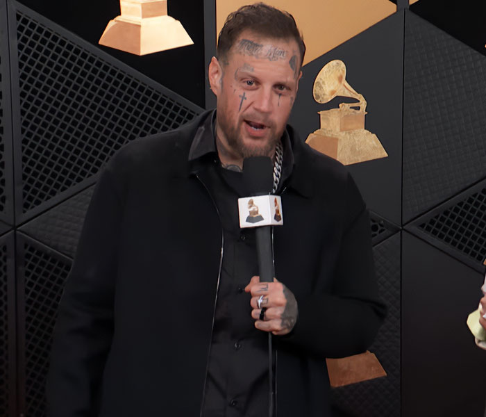 Male celebrity with face tattoos speaking into microphone against Grammy Awards backdrop during awkward celebrity reactions event.