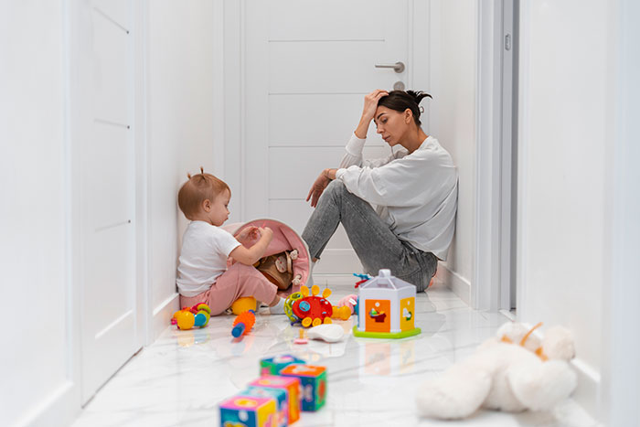 Tired mother sits on hallway floor near baby surrounded by toys, reflecting struggle with postpartum weight and pressure.