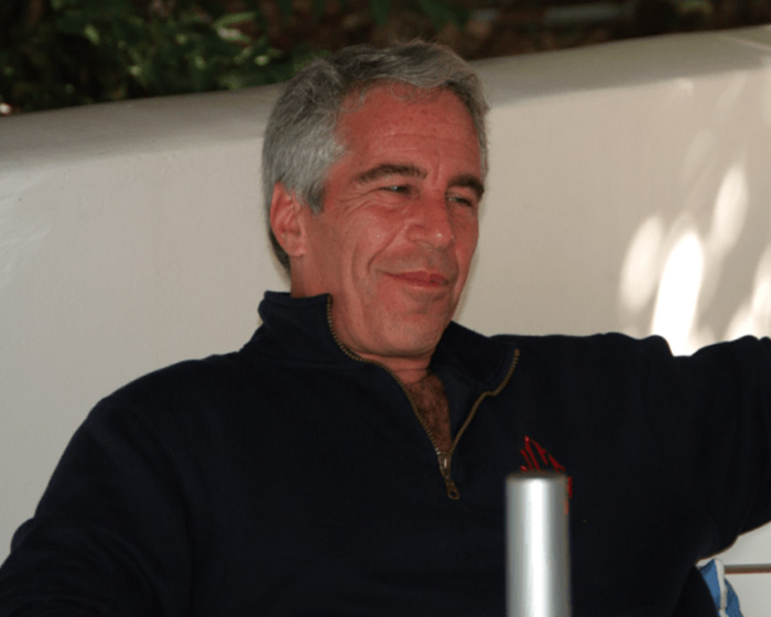 Man with gray hair wearing a navy jacket, seated and smiling in a casual indoor setting, related to Epstein files.