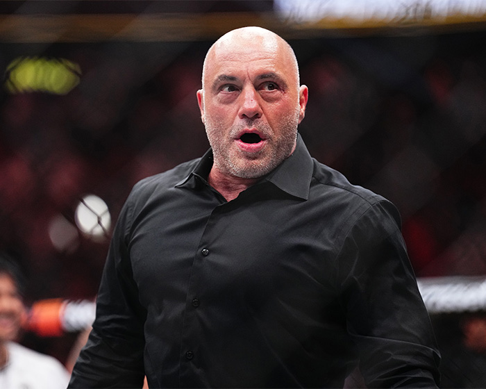 Joe Rogan in a black shirt inside an MMA cage reacting during a fight event with a blurred audience background.