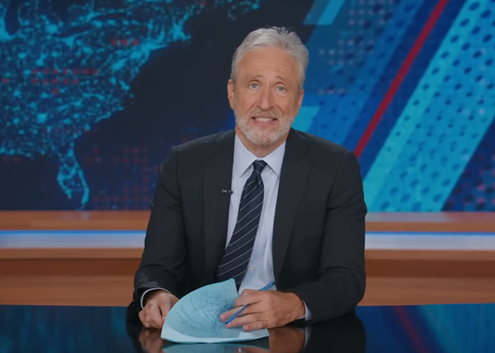 Jon Stewart seated at news desk in suit and tie, addressing the Epstein files in a televised broadcast.