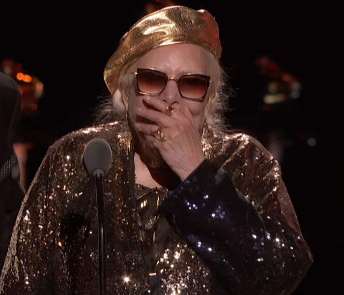 Elderly celebrity wearing gold sequined outfit and sunglasses, covering mouth in awkward reaction at Grammy Awards.