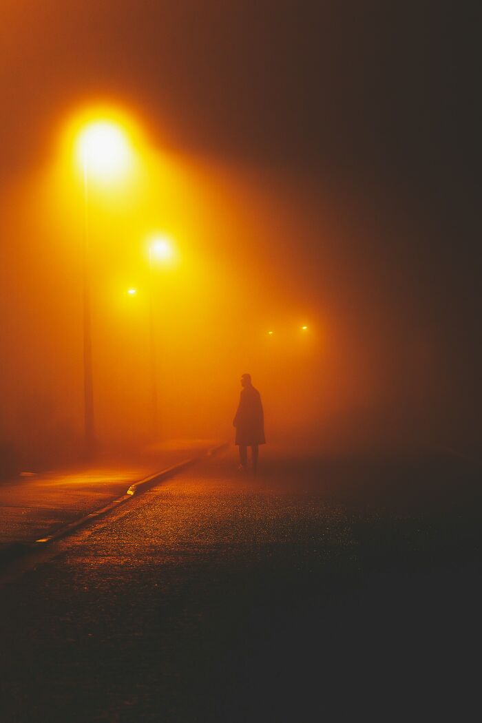 Silhouetted figure under orange streetlights on foggy night, evoking eerie graveyard shift worker atmosphere.
