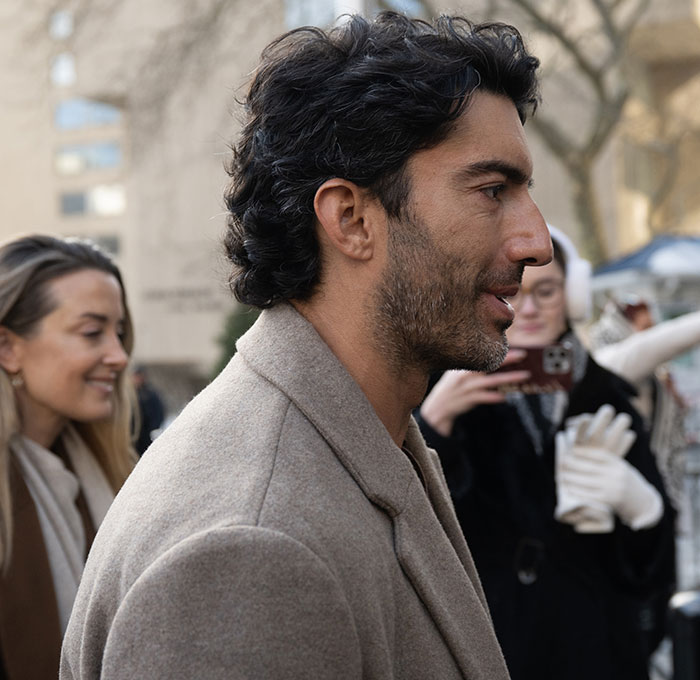 Man with dark curly hair and beard wearing a beige coat outdoors while people in the background record the moment.