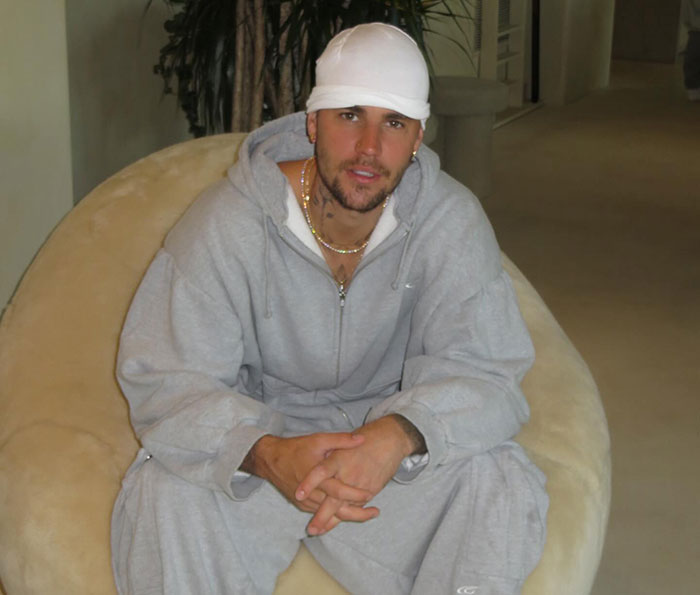 Justin Bieber wearing gray tracksuit and white cap sitting on a beige chair related to Epstein theory controversy.