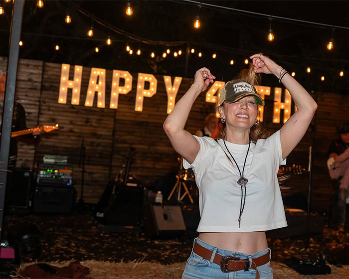 Woman smiling and lifting hair at party with festive lights and Happy 4th sign, reflecting on Botox moment on Big Bang Theory.