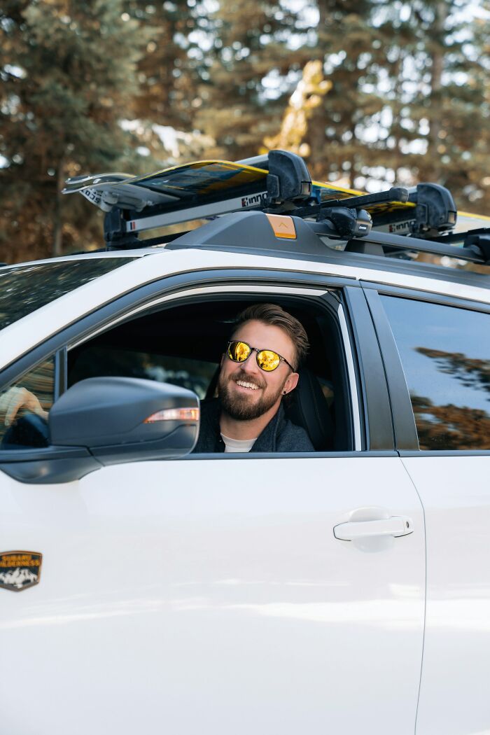 Smiling man wearing reflective sunglasses sitting in a white car with ski gear on the roof and trees in the background.