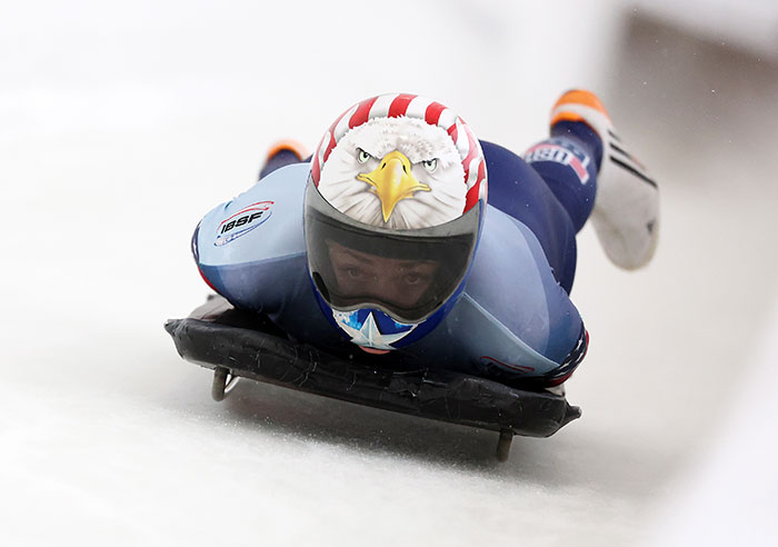 Olympic athlete in a skeleton racing suit with eagle helmet design speeding down an icy track at the Winter Olympics.