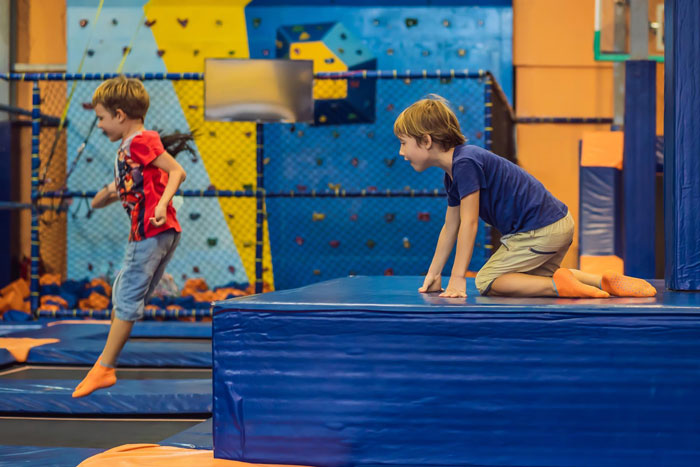 Two young boys playing and jumping at a trampoline park with colorful climbing walls in the background.
